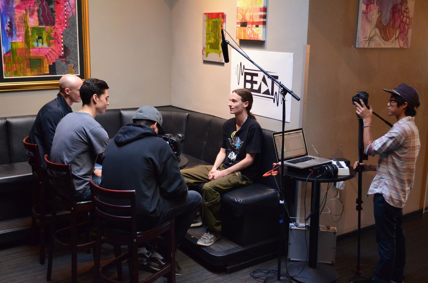 A contestant seated in front of the BEAT banner, talking to three people across from him, with a boom mic and two cameras in the room